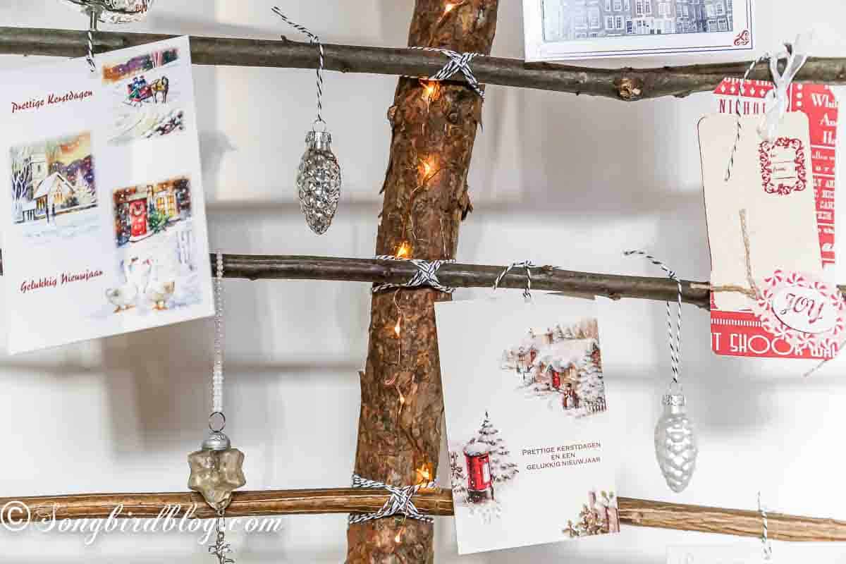 Creative ways to display Christmas cards. A rustic Christmas tree card holder made from natural branches. One big branch is standing in a tray and six horizontal branches are attached with twine. Christmas cards are hanging on the branches.