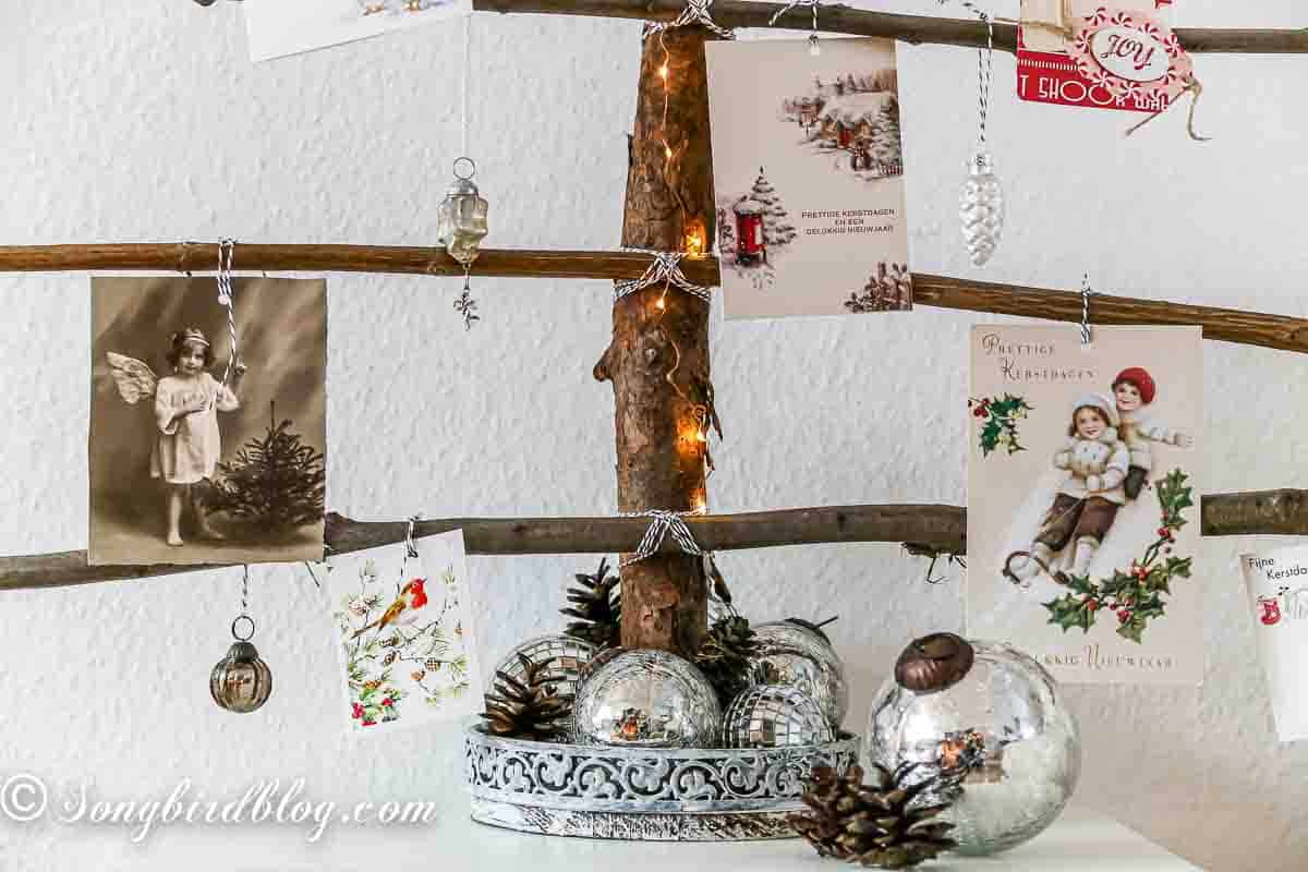 Creative ways to display Christmas cards. A rustic Christmas tree card holder made from natural branches. One big branch is standing in a tray and six horizontal branches are attached with twine. Christmas cards are hanging on the branches.