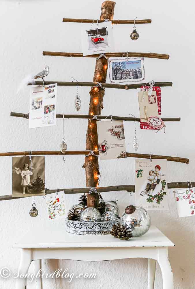 Creative ways to display Christmas cards. A rustic Christmas tree card holder made from natural branches. One big branch is standing in a tray and six horizontal branches are attached with twine. Christmas cards are hanging on the branches.