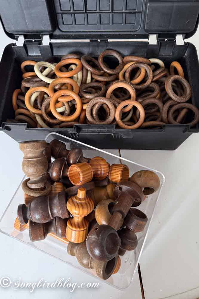 Black tool box filled with various wood curtain rings beside a clear box with wooden curtain finials.