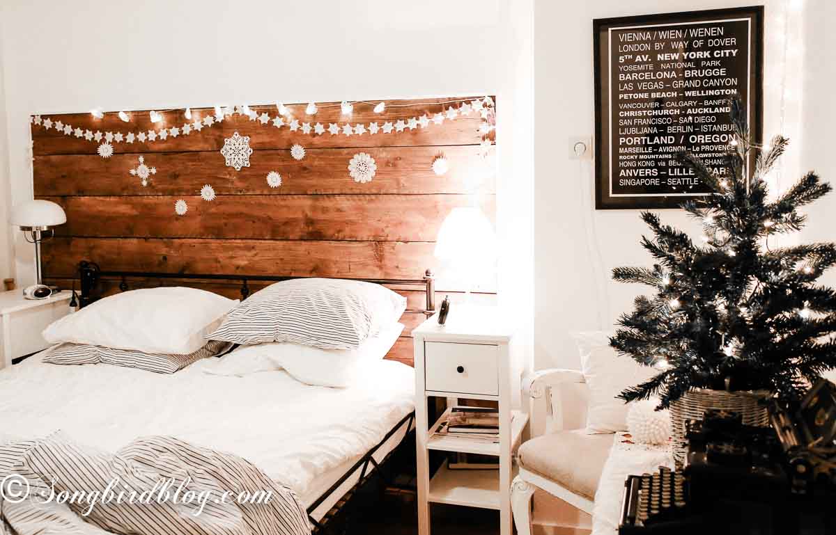Whole room view of a simple Christmas bedroom at night, lit by the glow of a small tree with lights only and a string of lights draped over the headboard.