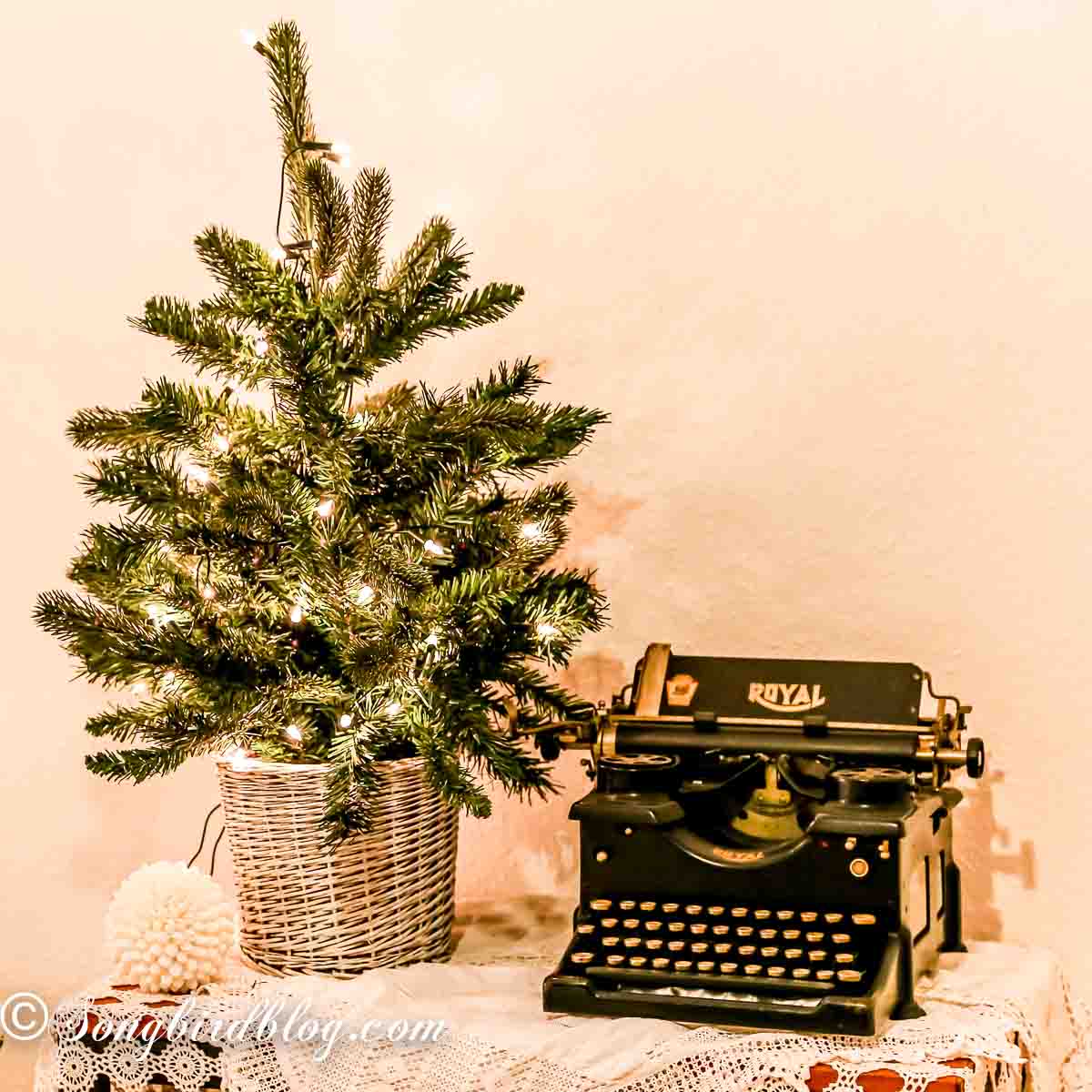 Vintage linens draped over an old wooden table. On top a small Christmas tree with lights and a vintage typewriter.