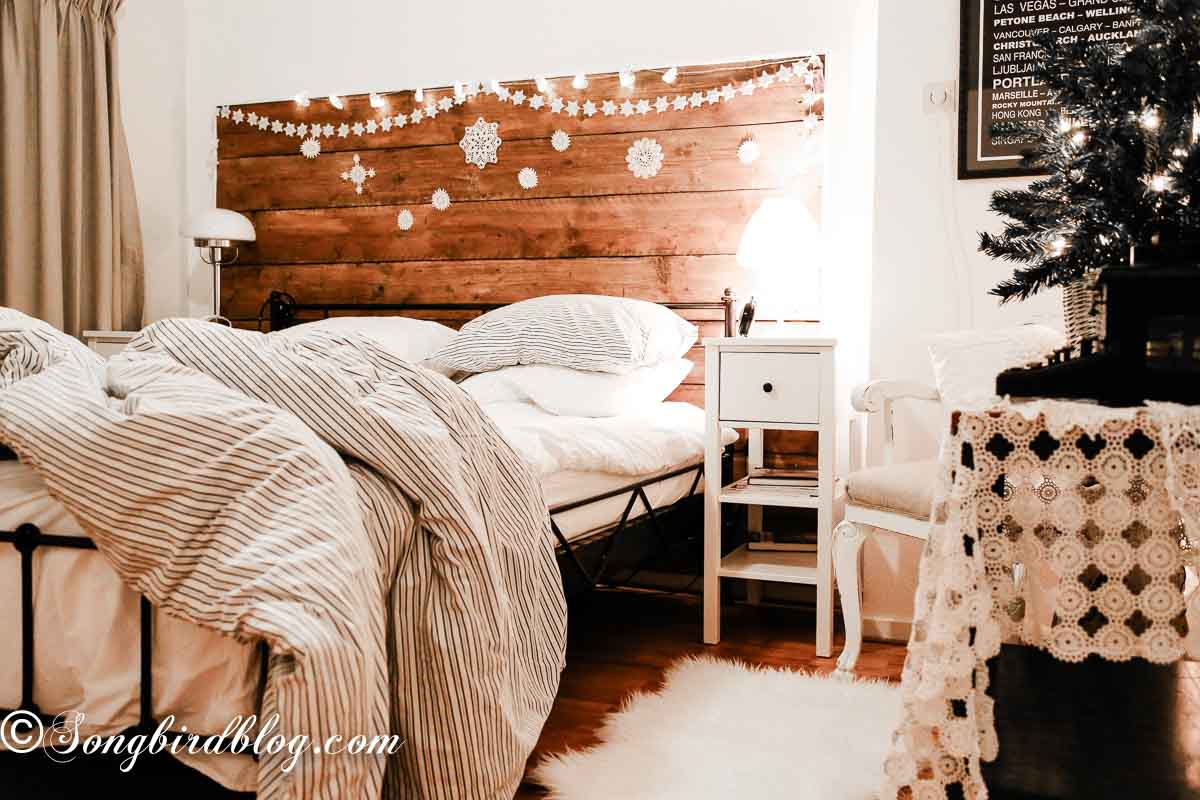 Cozy Christmas bedroom seen from the doorway, with a small tree's lights reflecting off vintage linens and soft garland lights above the bed.