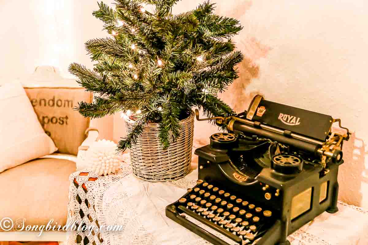 An old table dressed with vintage linens. On top a small Christmas tree with lights and a vintage typewriter.