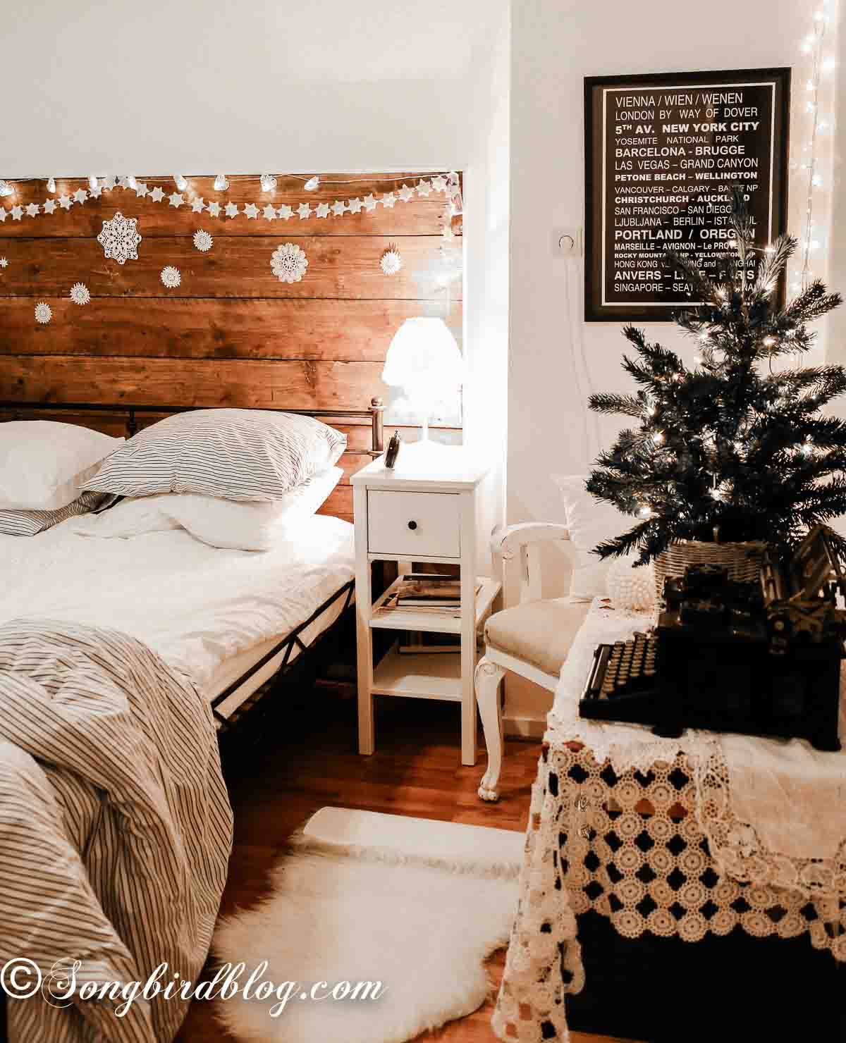 Wide view of a cozy Christmas bedroom with a small lit tree on a vintage linen covered table, a garland and doilies on the dark wood headboard, and warm light glowing throughout.