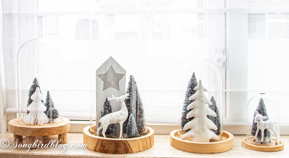 Row of four glass cloches filled with Christmas decor on a window sill.