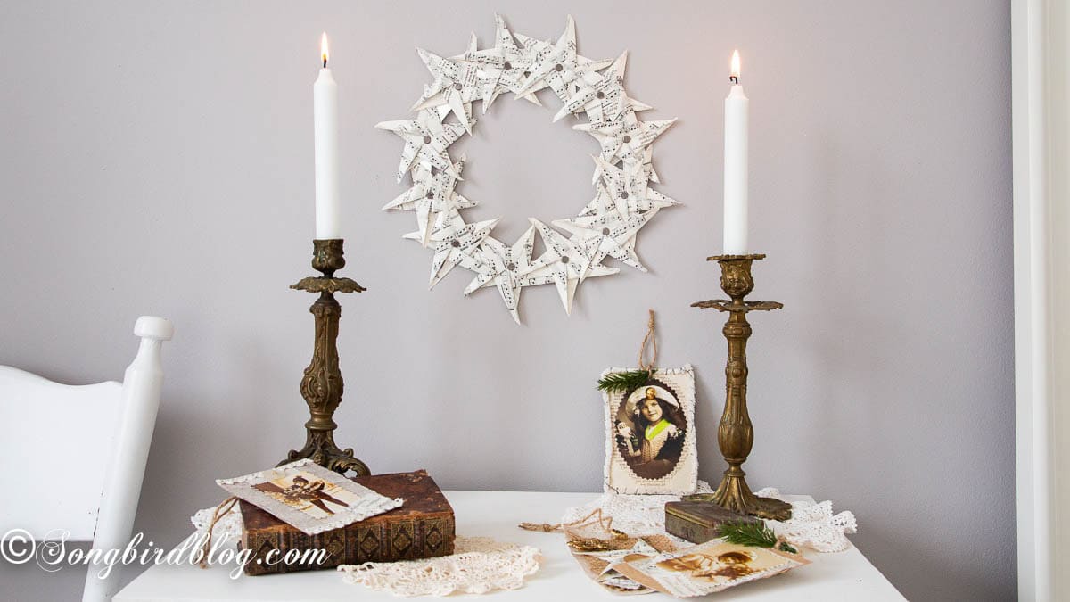 Paper star wreath made from vintage sheet music pages hanging on a wall above a white sidetable styled with antique books, candles, and trinkets.