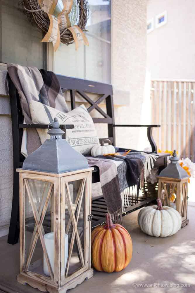 Farmhouse style front porch decorated for fall with a bench with blankets and pillow, two lanterns and faux pumpkins.