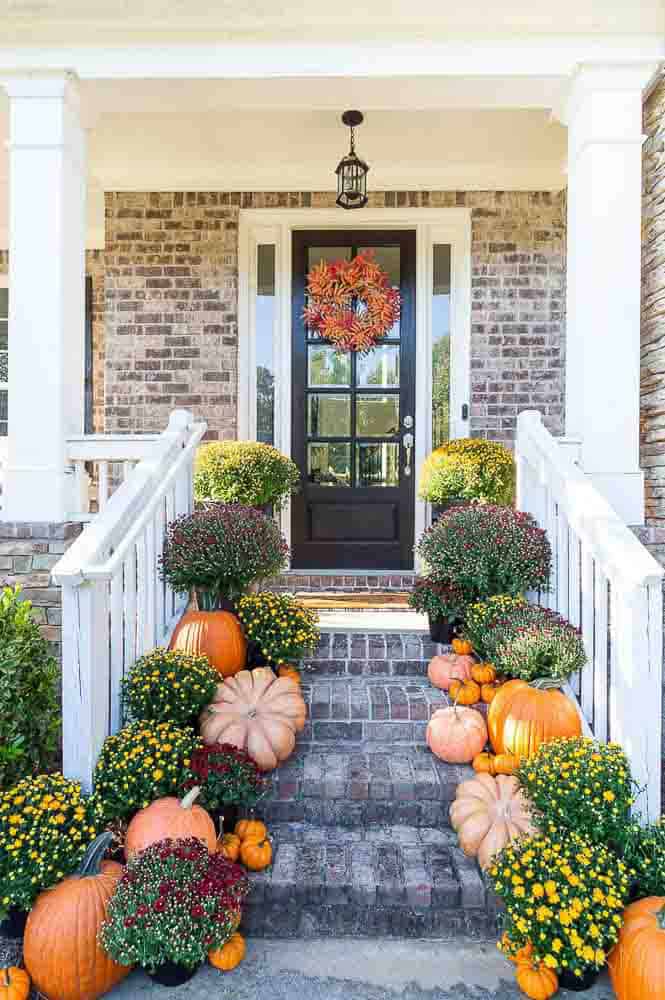 Fall entryway with orange pumpkins and yellow mums lined up along the steps leading to a dark front door with a colorful fall wreath.