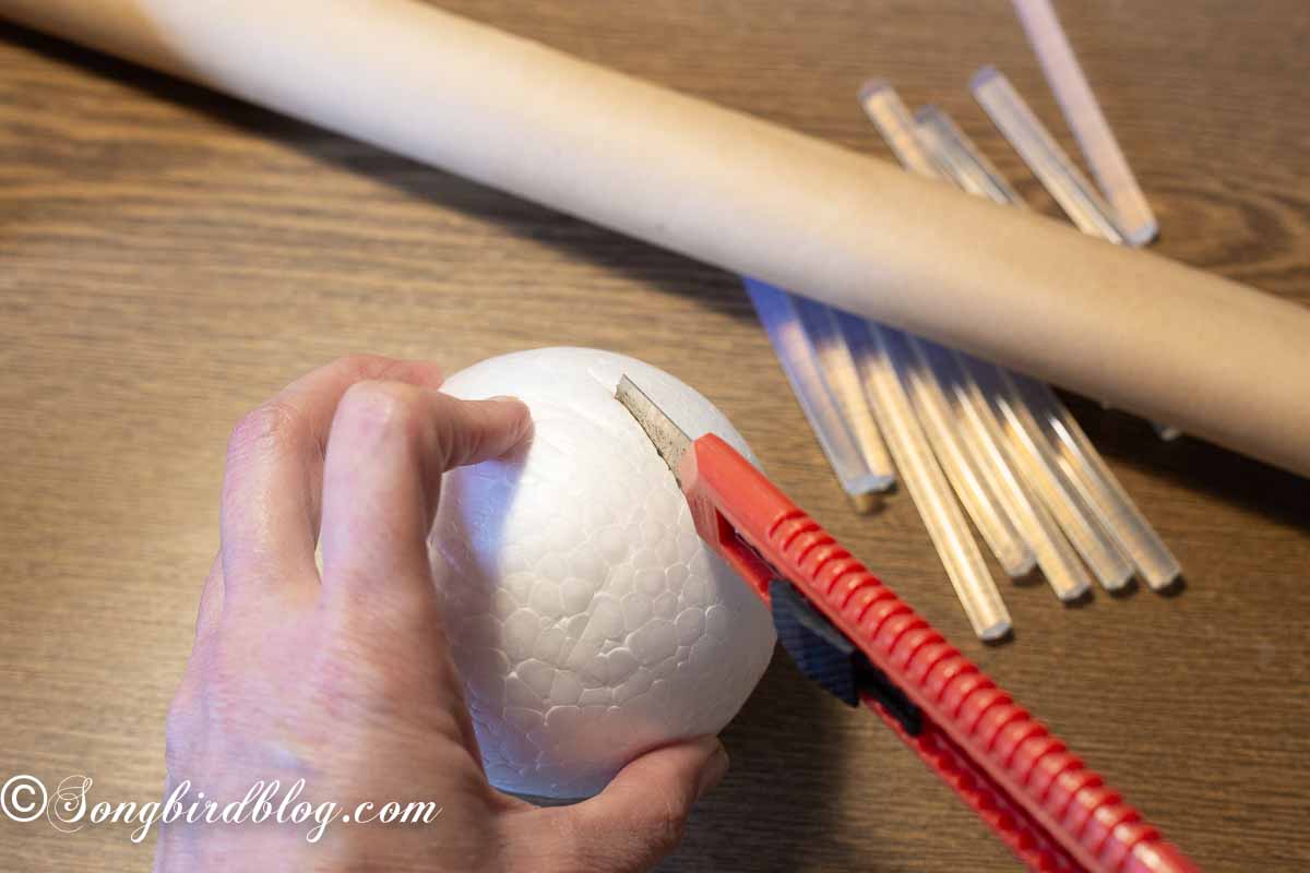 A hand holding a styrofoam ball and an exacto knife cutting the bottom part of the ball.