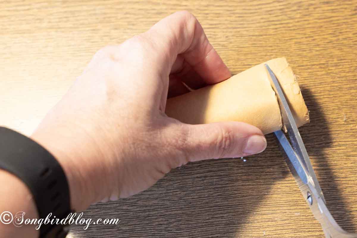 A hand holding a role of brown craft paper and scissors cutting a roll of half an inch of the top.
