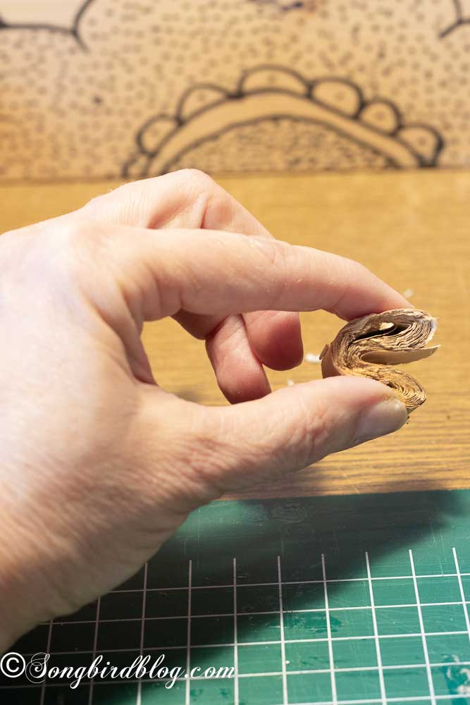 Two fingers pinching a roll of brown craft paper into a heart shape.