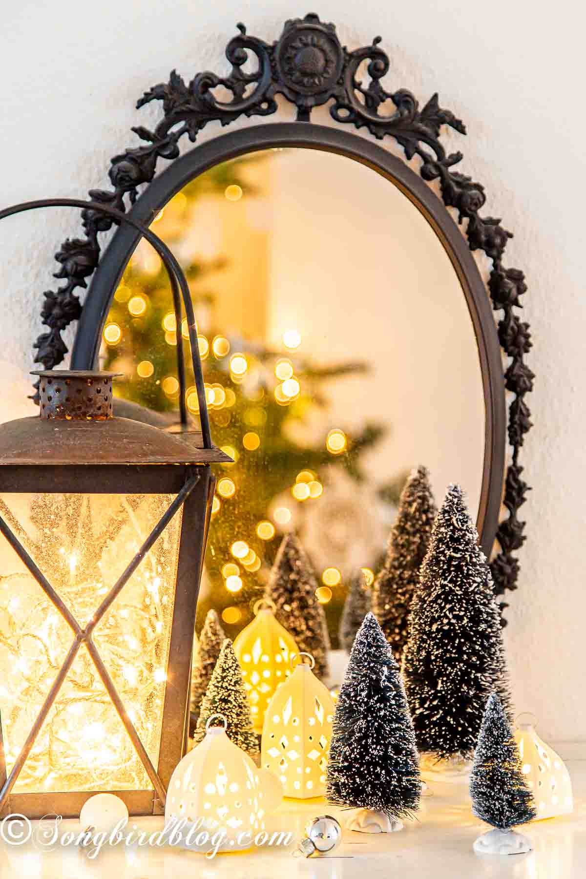 A simple Christmas display with an outdoor lantern filled with Christmas lights, bottle brush trees and little white candle holders in front of a mirror.