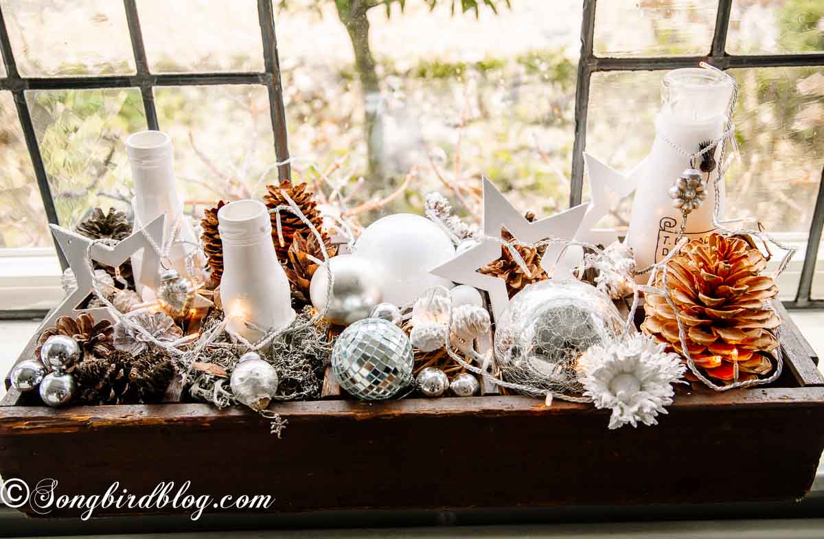 Wooden crate on a window sill filled with ornaments, pinecones and a string of christmas lights.