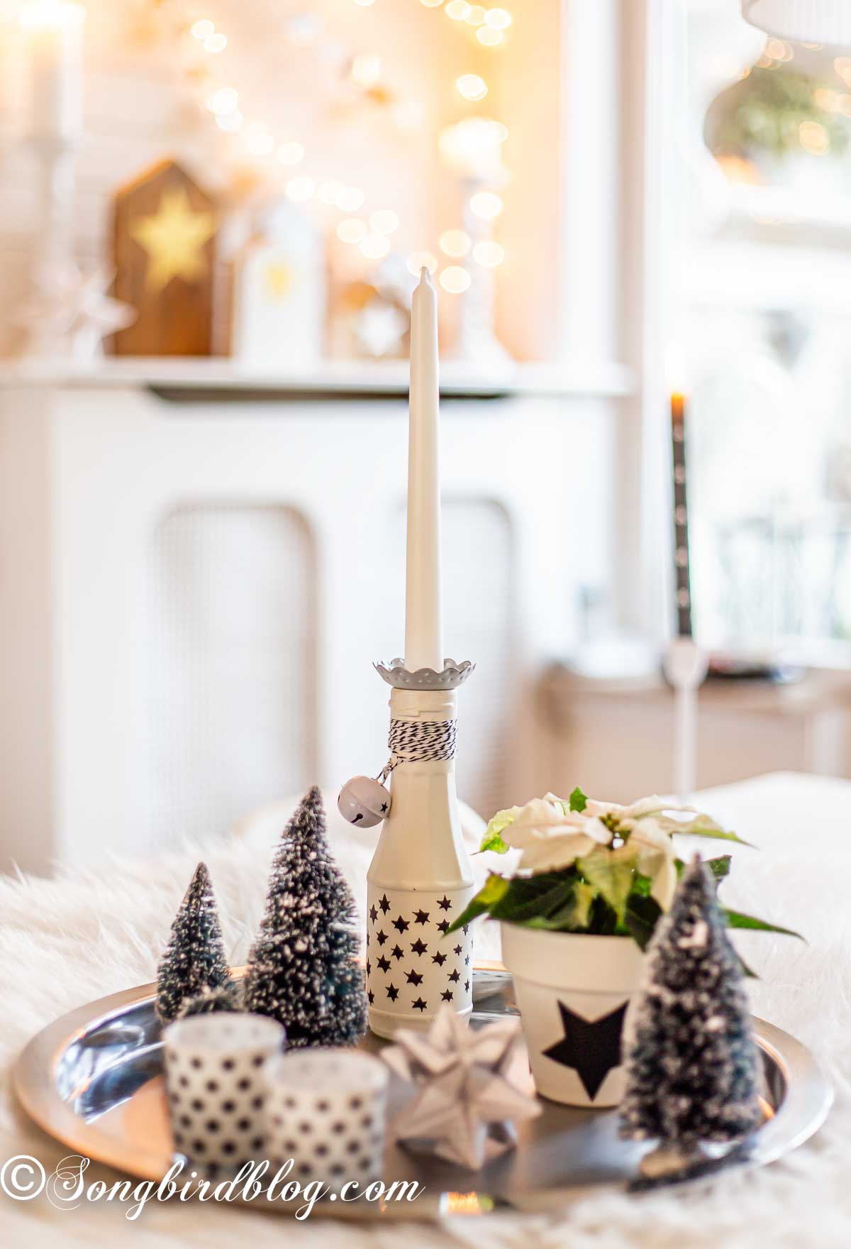 A Christmas vignette on a tray with bottle brush trees, black and white candle holders with stars and a white painted bottle with a candle. Christmas lights on a mantel in the background.