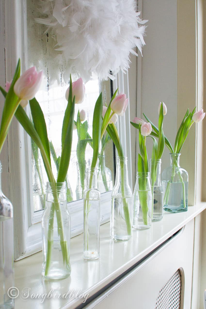 Pink tulips in vintage bottles lined up in front of a mirror on a mantel fireplace.