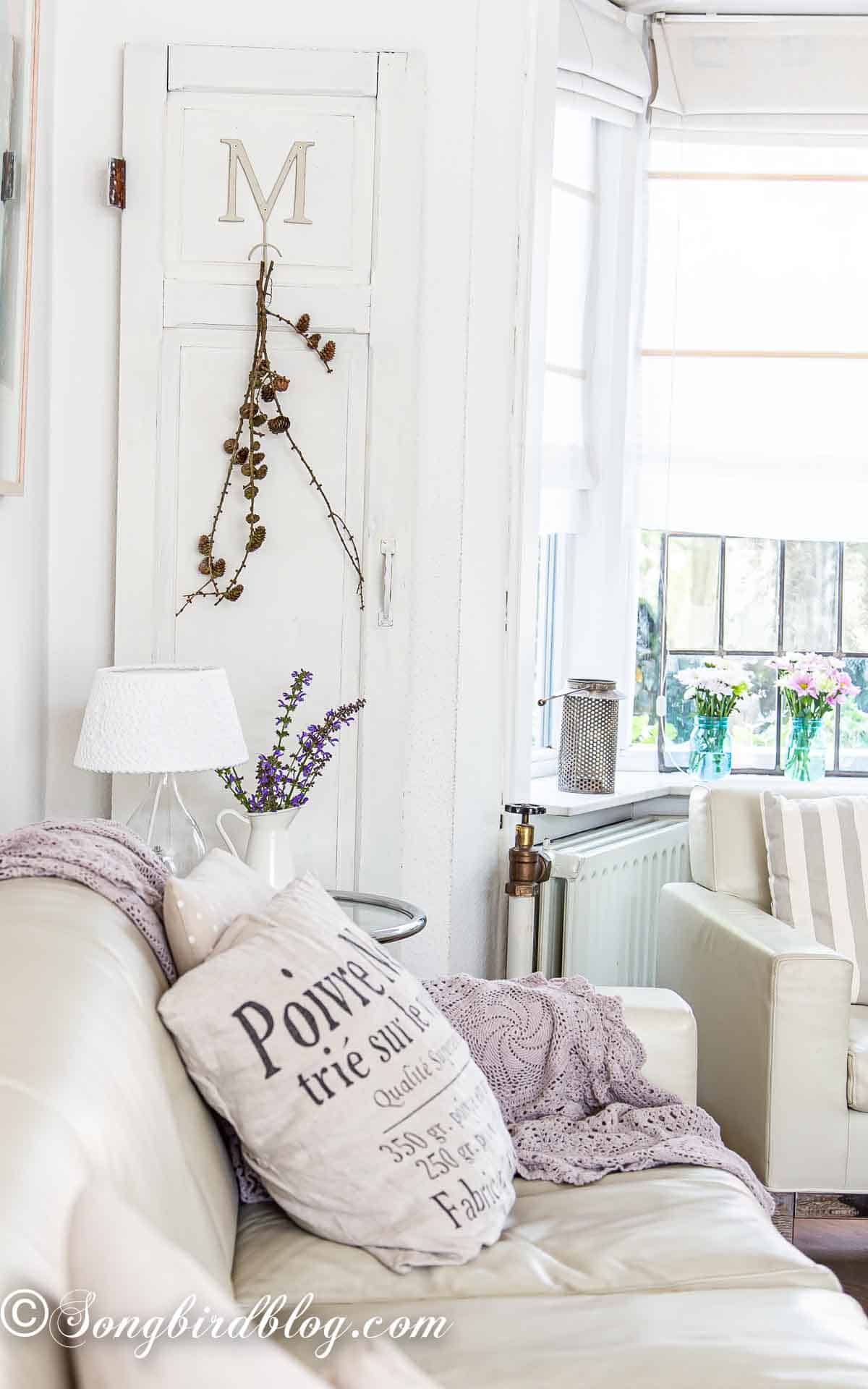 Living room with couch and pillows and a leaning door against the wall. Braches with small pinecones are hanging from a hook on the door.