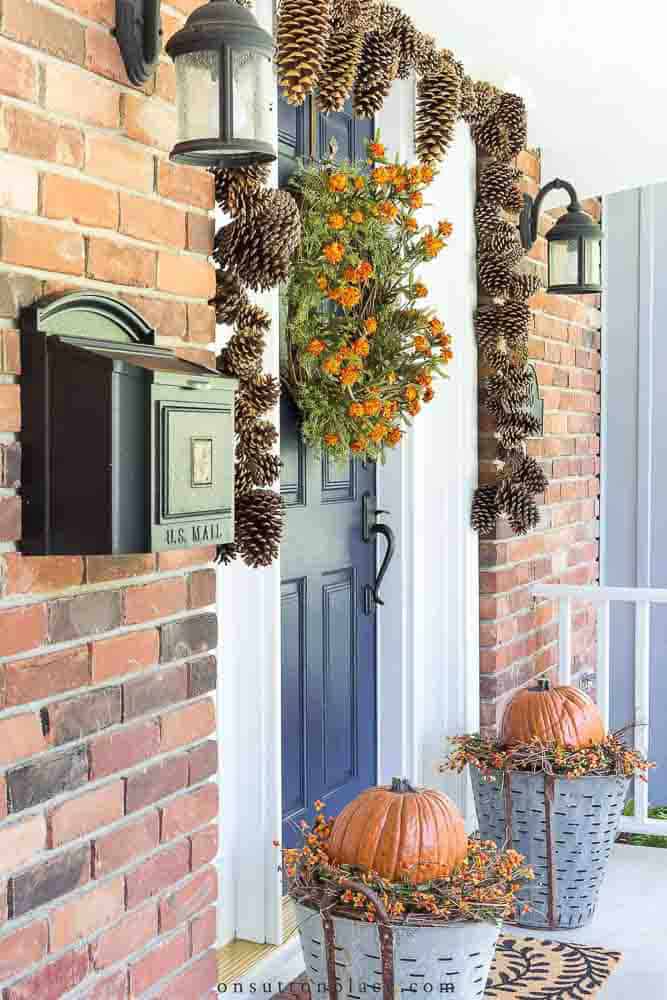 Front door with a orange flower wreath and a pinecone garland around the frame. Flanked by two planters made from olive baskets and filled with pumpkins.
