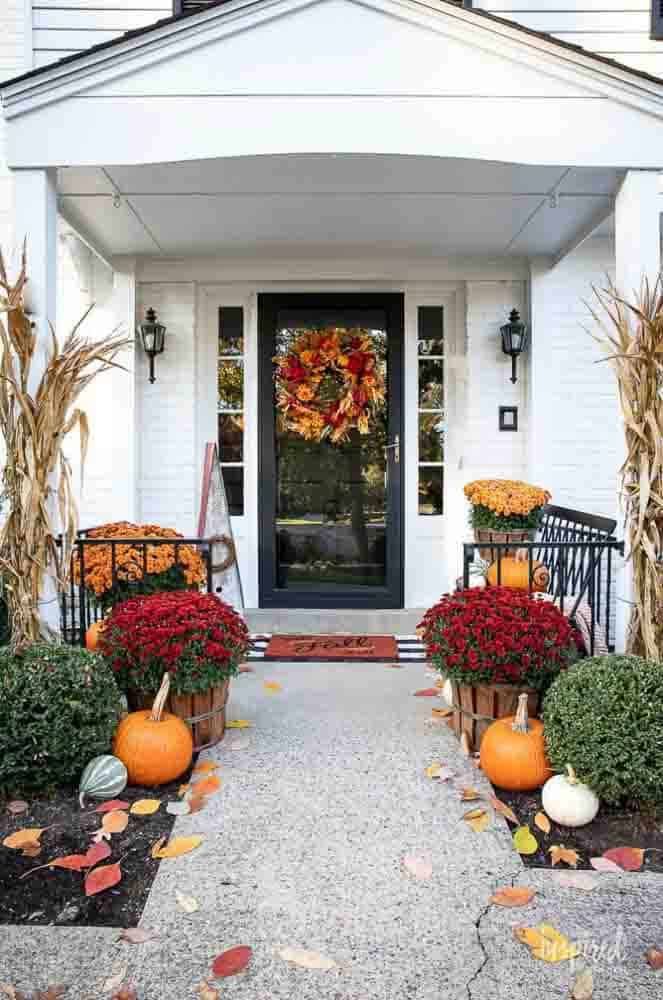 White entryway decorated for fall with red and orange pumpkins, mums and a fall wreath.