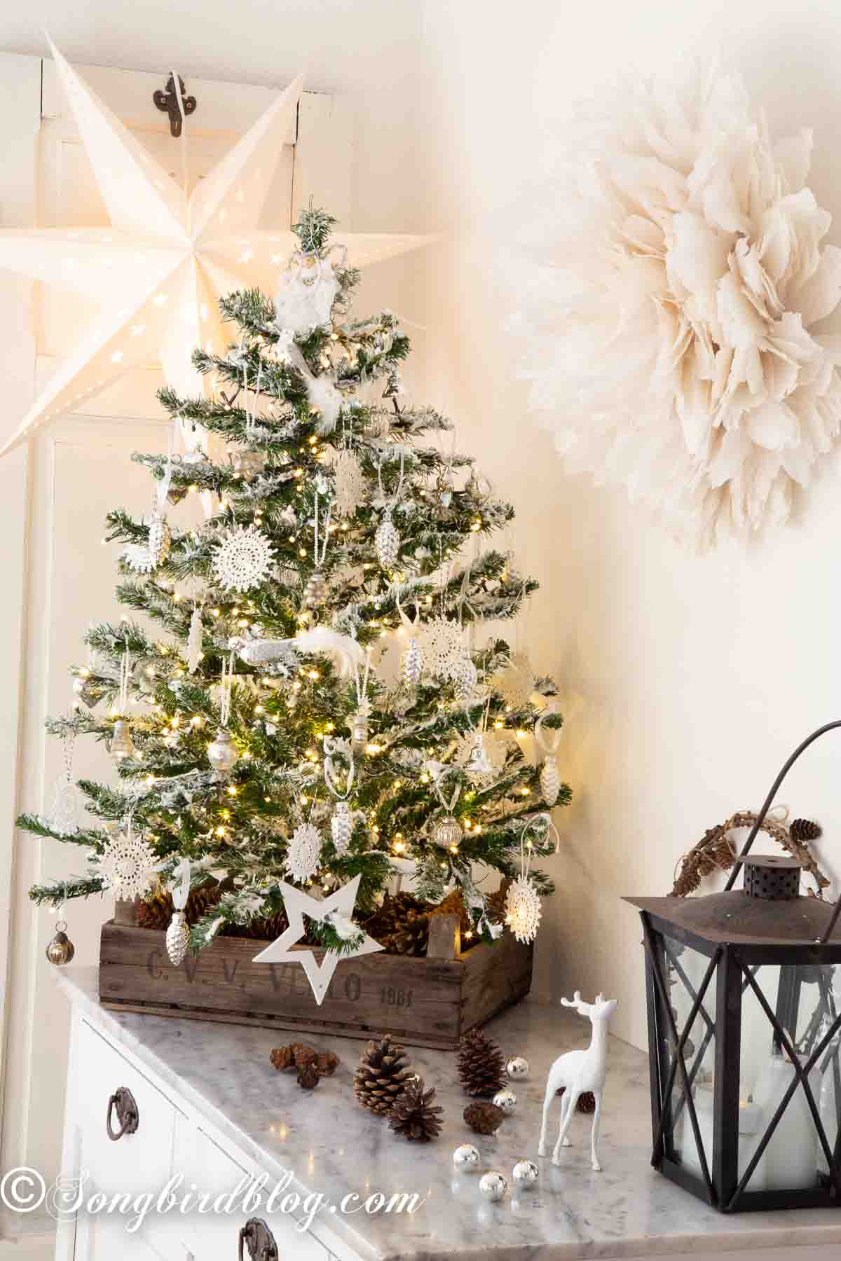 A small flocked Christmas tree in a crate on a sideboard with a lantern, deer figurine and pinecones.