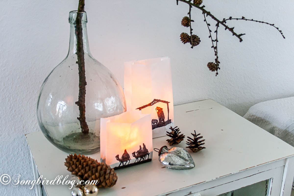 Two hand-traced paper bag lanterns lit by battery candles, arranged with pinecones and silver Christmas ornaments on a table.