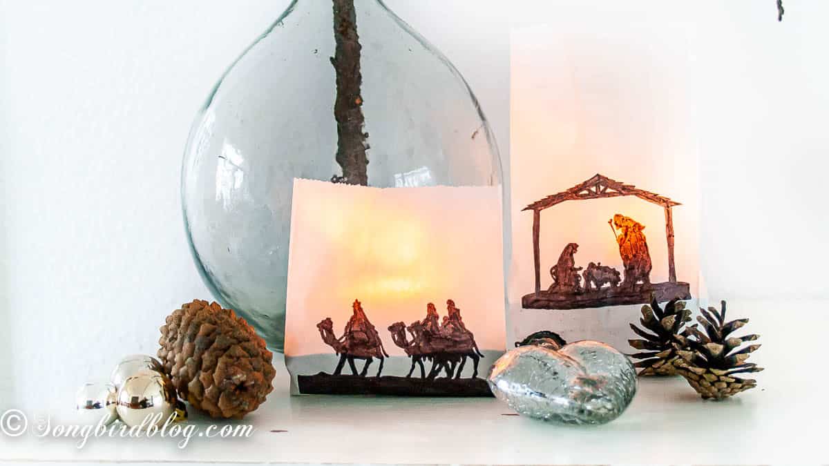 Two lit nativity paper bag luminaries, one with a stable and Mary and Joseph silhouette, the other with the three kings, in front of a clear vase of tall pinecone branches with scattered pinecones and silver ornaments.