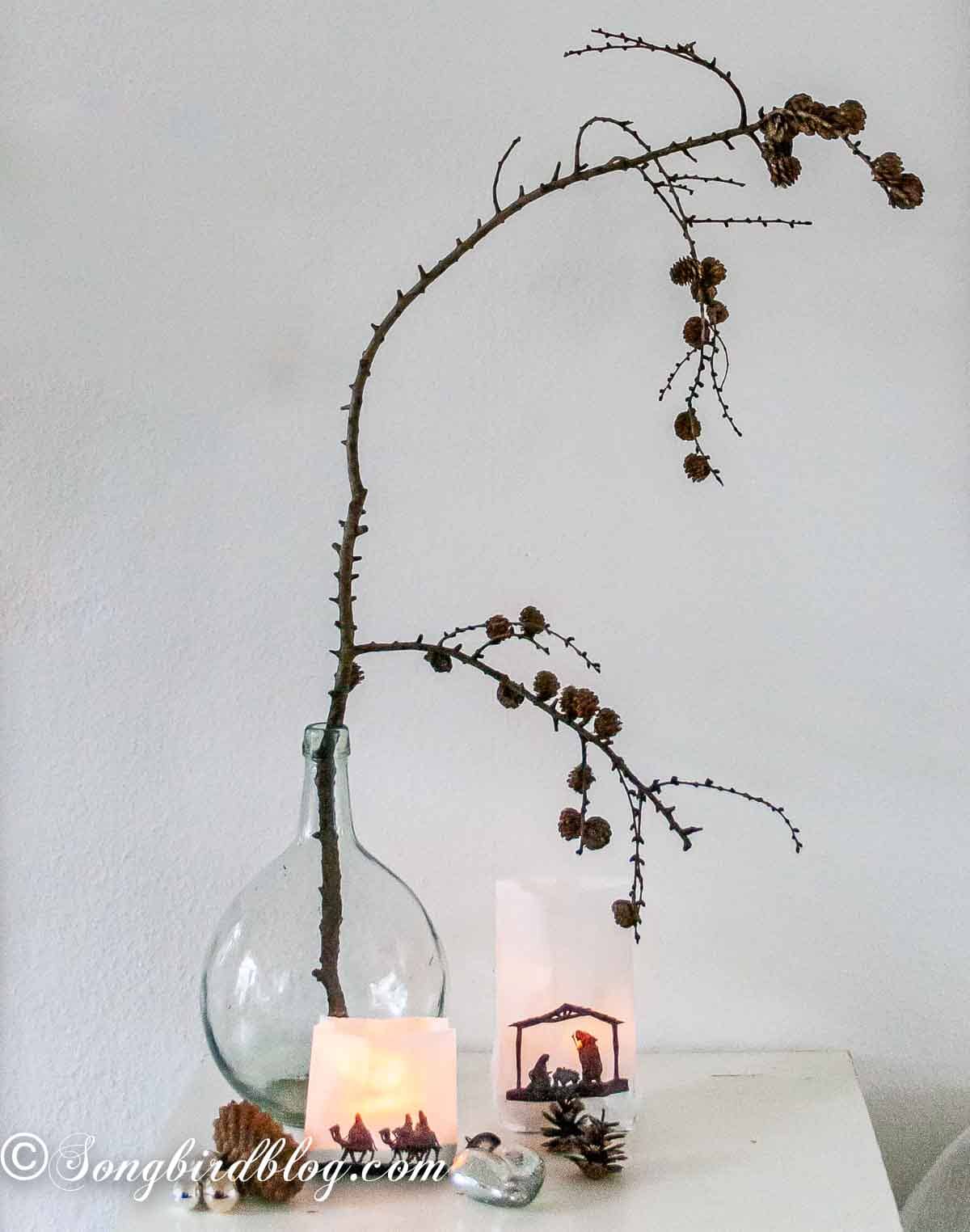 A Christmas display of glowing handmade luminaries beside a clear vase filled with pinecone branches and silver ornaments.