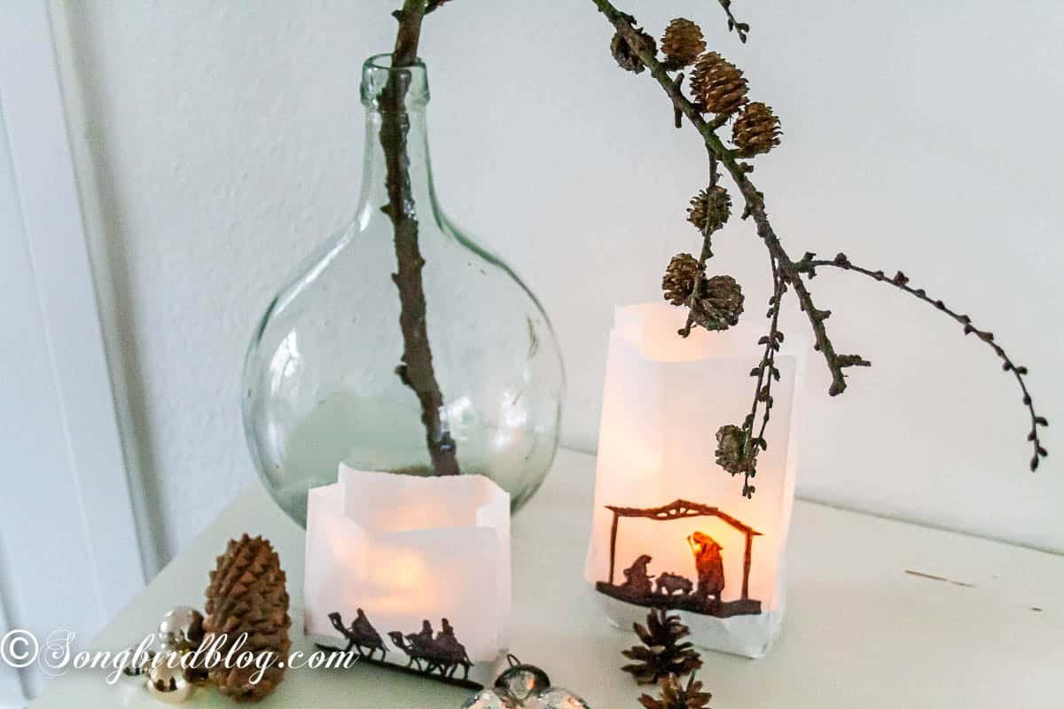 Nativity paper bag luminaries glowing in a Christmas vignette with a tall clear vase of pinecone branches, silver ornaments, and pinecones.