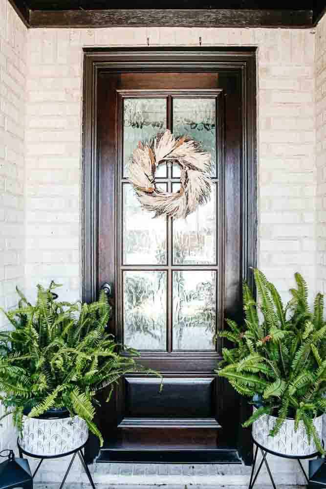 Wheat wreath on a brown front door flanked by two ferns in planters.