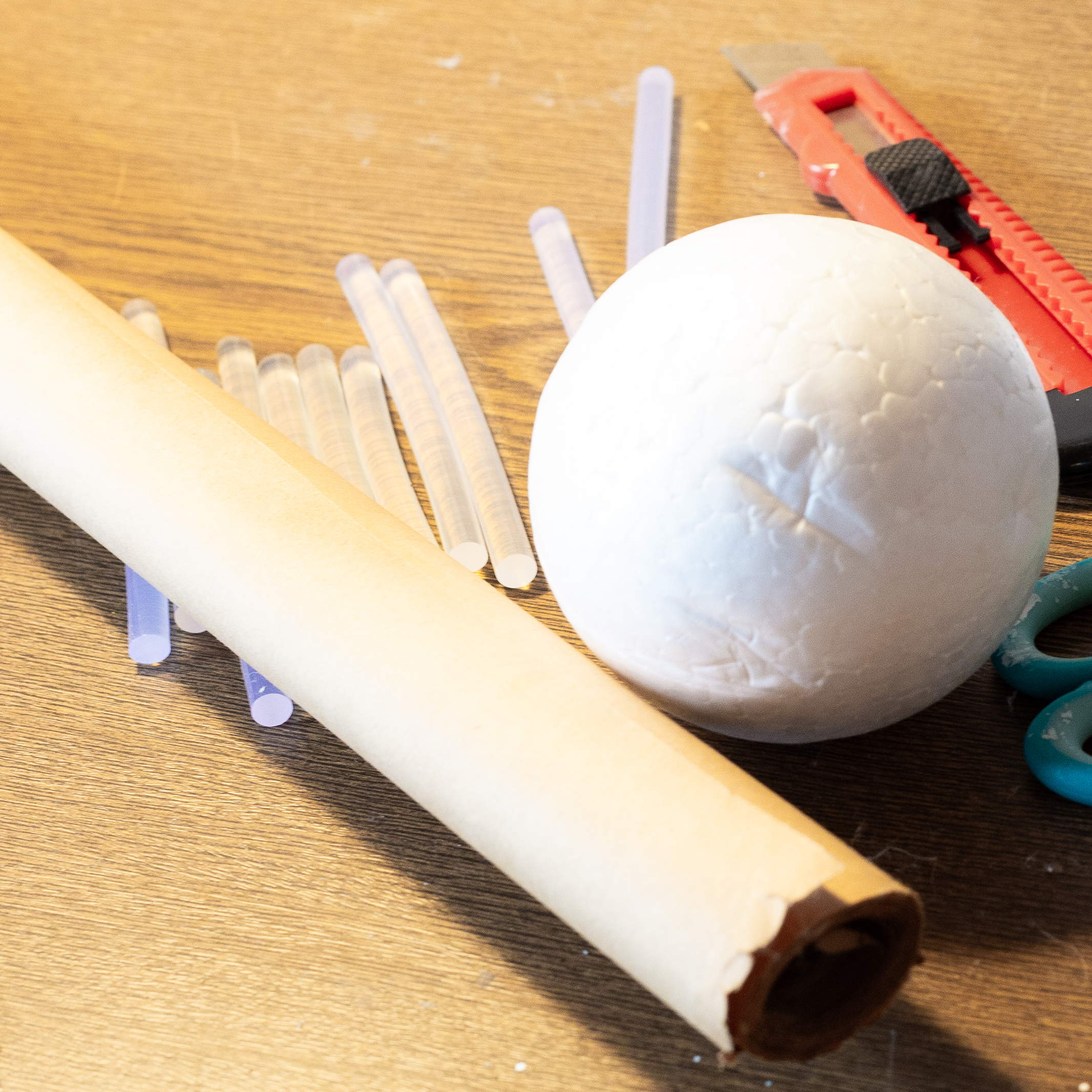 A grouping of a white styrofoam ball, a roll of brown craft paper, small glue sticks and an exacto knife.