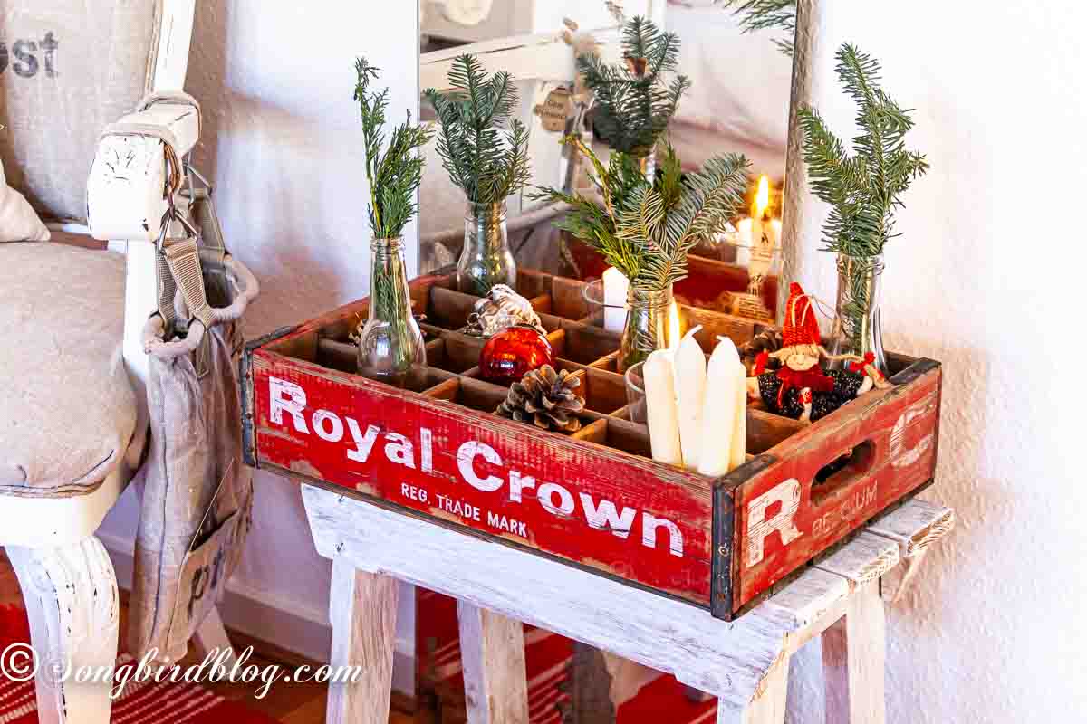 A red crate on a stool, filled with Christmas ornaments, candles and greenery.