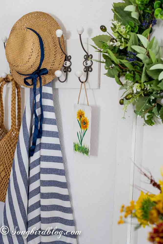 A shelf with hooks featuring a hand painted card with sunflowers, a straw summer hat and a blue and white striped towel.